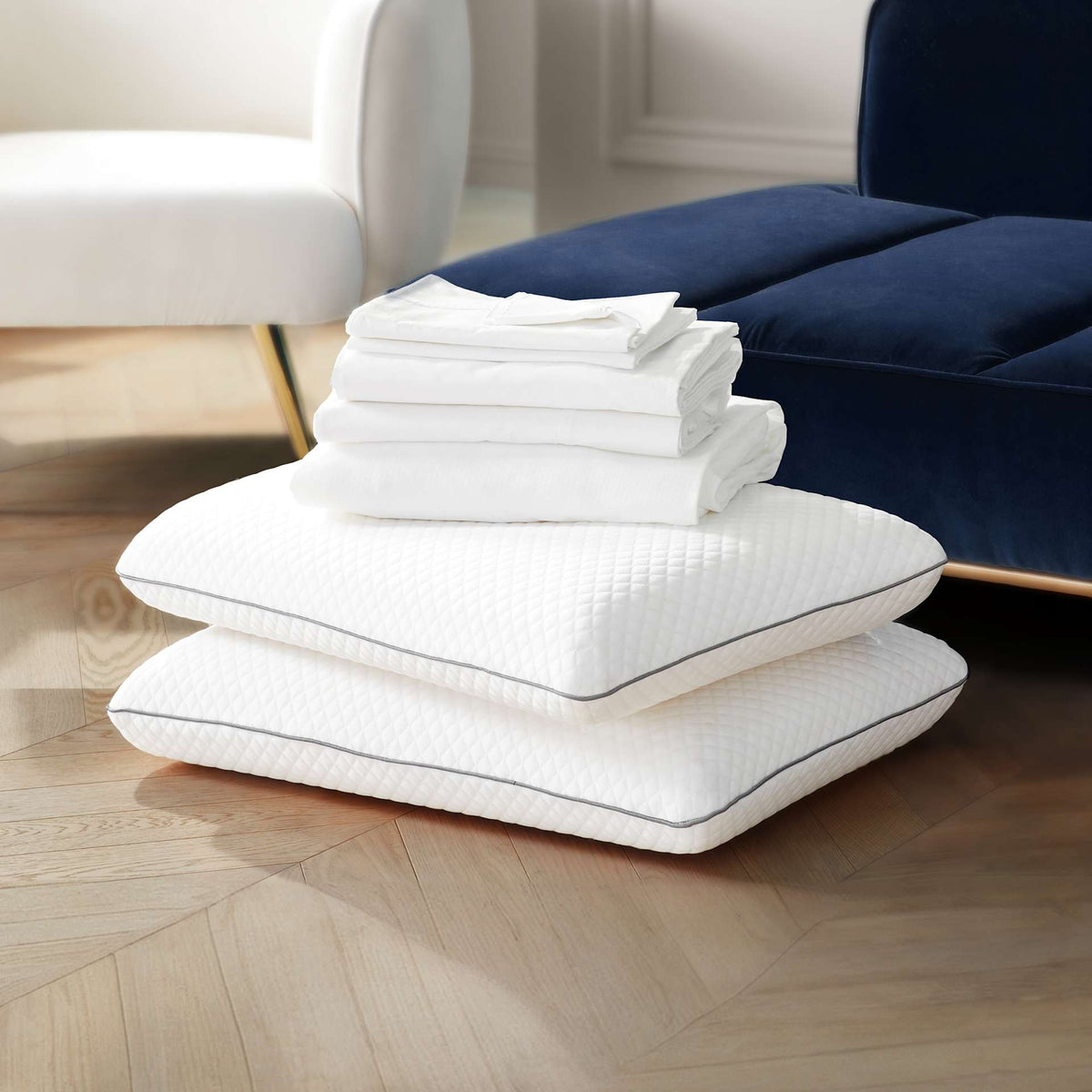 Two white quilted pillows stacked on a light wood floor, with a folded stack of white bedding or towels placed on top, in a softly styled room with a white chair and a navy sofa in the background.