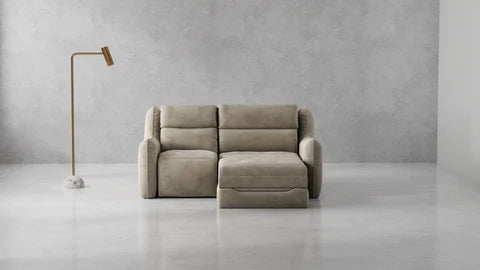 A modern beige sectional sofa with a chaise lounge, set in a minimalist room with a textured gray concrete wall and floor, next to a slim floor lamp.