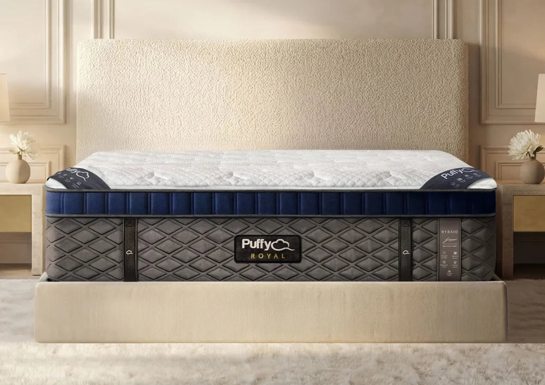 A Puffy Royal mattress shown on a bed in a styled bedroom setting. The mattress features a white quilted top, a navy blue side bolster, and a dark gray quilted base with the Puffy Royal logo on the front.