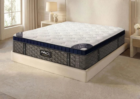 A mattress on a low platform bed in a softly lit bedroom. The mattress has a white quilted top and dark gray patterned sides with navy trim and visible Puffy brand labels.